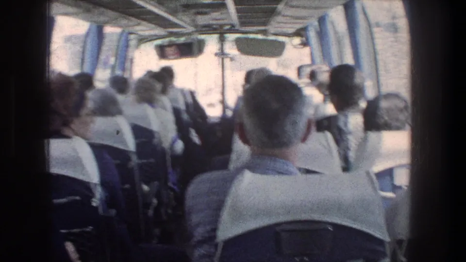 1976 Jerusalem home-movie frame showing passengers seated inside a bus, seen from the rear with blue curtains and bright windows.