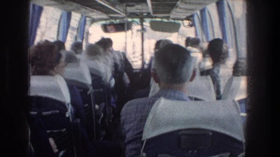 1976 travel footage frame looking down the bus aisle toward passengers and windows with pale headrest covers.