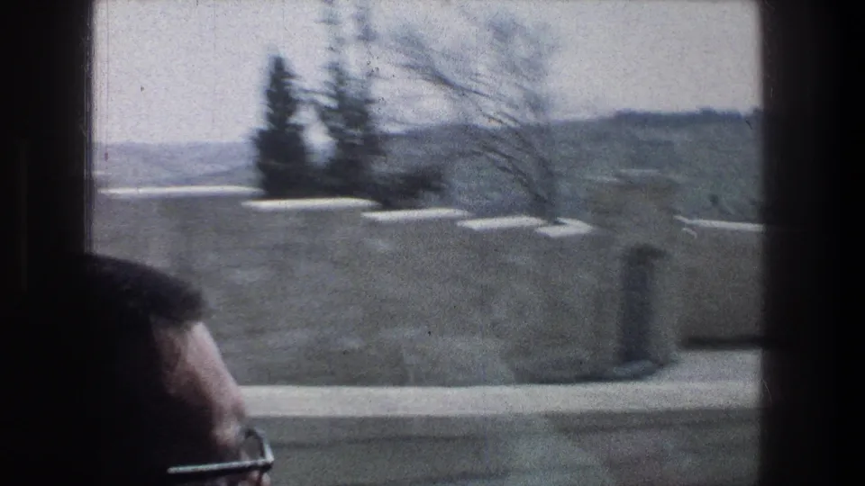1976 Jerusalem home-movie frame showing a passing view through a vehicle window, pale stone wall, distant hills, and a passenger head in the foreground.