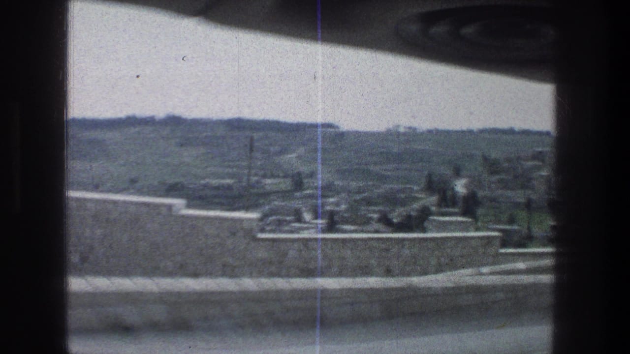 1976 Jerusalem frame looking through a moving vehicle window toward a pale stone wall, low terrace edge, scattered figures, and hilly landscape.