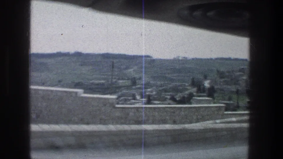 1976 Jerusalem frame looking through a moving vehicle window toward a pale stone wall, low terrace edge, scattered figures, and hilly landscape.