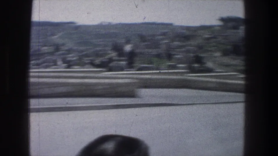 1976 Jerusalem 8mm frame with motion blur from a vehicle, showing a road or terrace edge, pale wall, trees, and distant hillside.