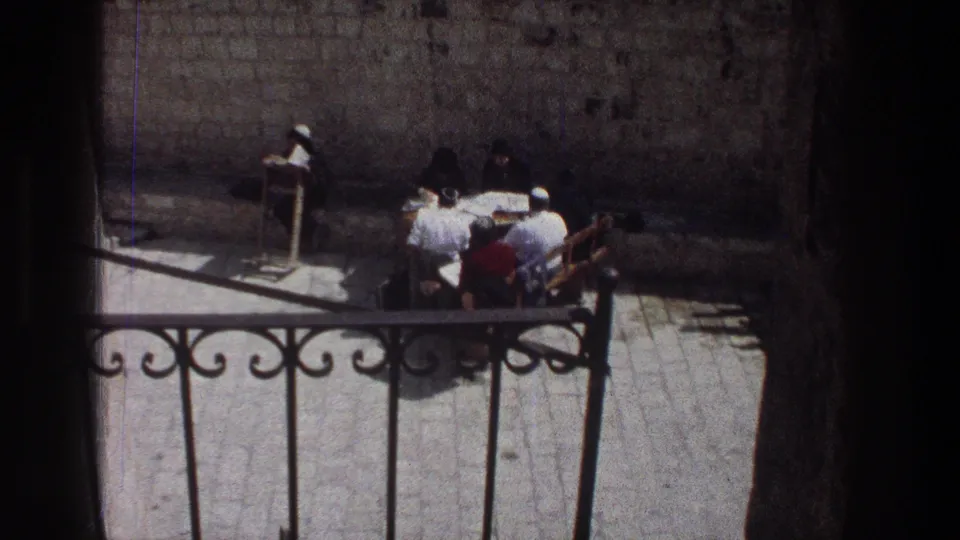 1976 Jerusalem home-movie frame showing people seated or standing around a courtyard table behind decorative iron railing and a stone wall.