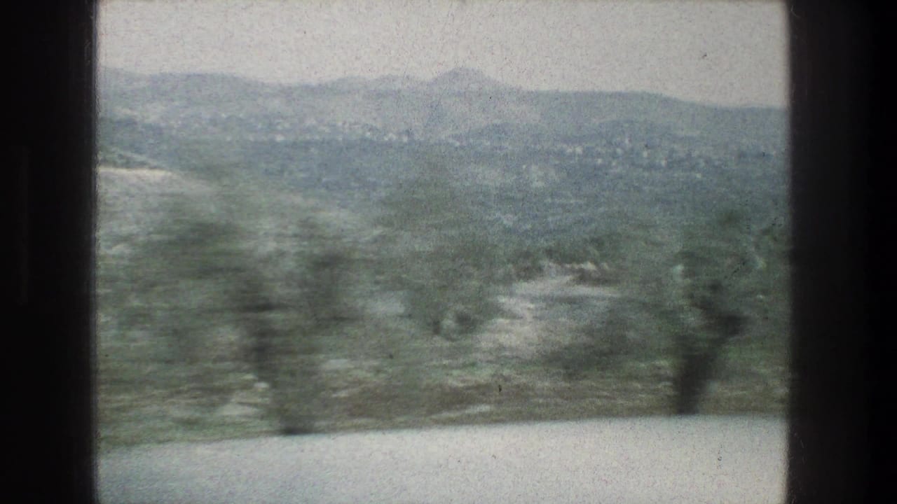 Hazy 1976 8mm view near Jerusalem with dark window dividers, rolling hills, scrub vegetation, and blurred foreground trees.