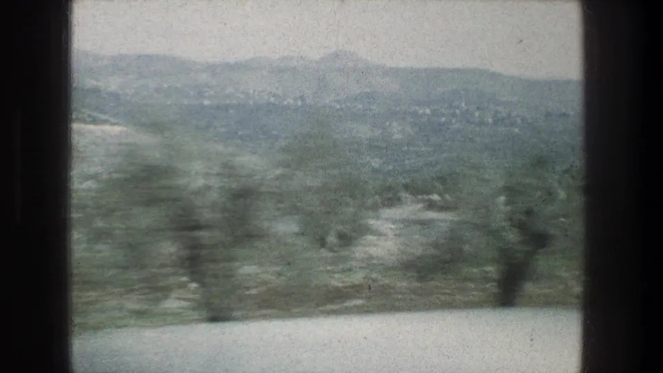 Hazy 1976 8mm view near Jerusalem with dark window dividers, rolling hills, scrub vegetation, and blurred foreground trees.
