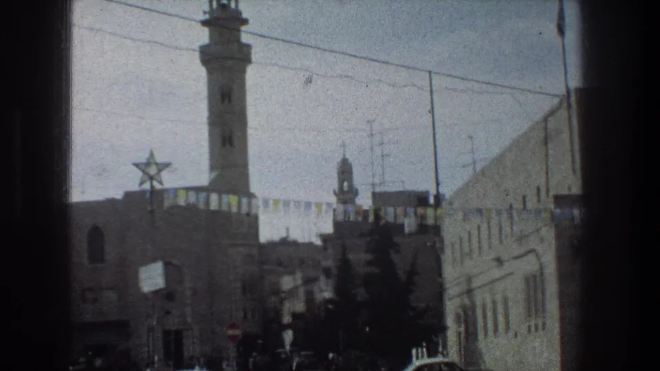 1976 Jerusalem street seen from a moving window, with a minaret, Star of David decoration, bunting, stone buildings, and trees.