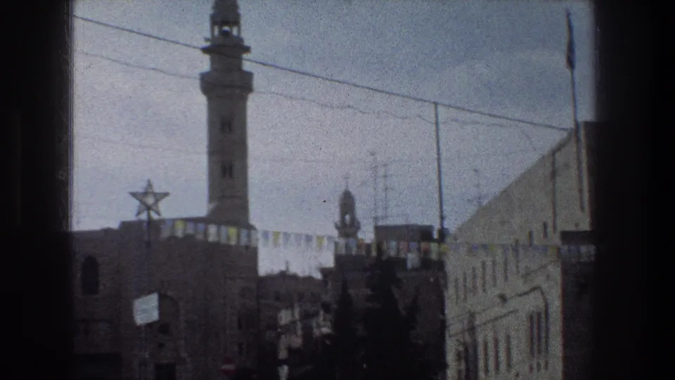 1976 Jerusalem home-movie frame showing a minaret above a decorated street with a Star of David, bunting, buildings, trees, and a traffic sign.