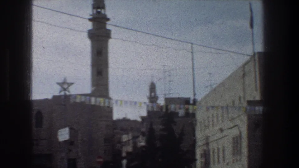 1976 Jerusalem street scene from a moving train or vehicle, with soft focus on the minaret, Star of David decoration, bunting, buildings, trees, and a car edge.