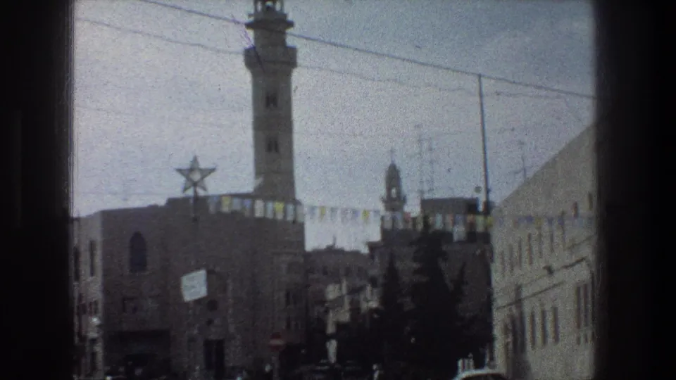 Jerusalem street frame from 1976 with a tall minaret, Star of David decoration, bunting across the road, trees, and stone facades.