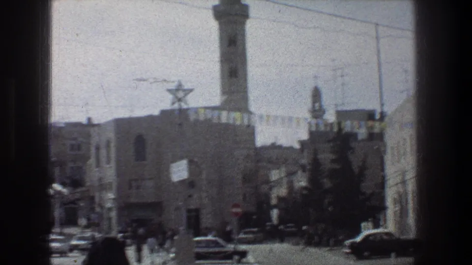 1976 Jerusalem view with a minaret partly high in frame, Star of David decoration, classic cars, distant pedestrians, bunting, and stone buildings.