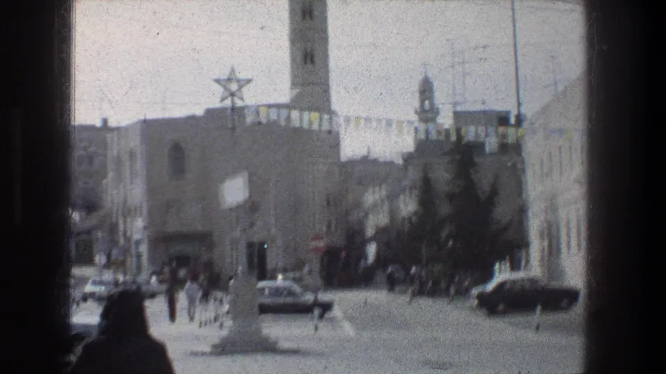 Late frame of the 1976 Jerusalem street view showing cars, distant pedestrians, a minaret, Star of David decoration, overhead wires, and bunting.