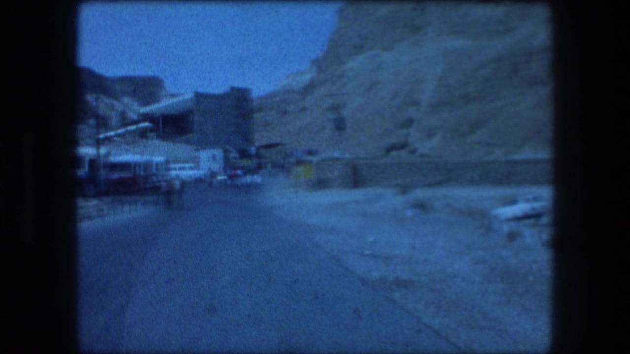 Vehicle-view frame in Messada, Israel, showing a road, roadside structures, rocky mountains, and clear blue sky.