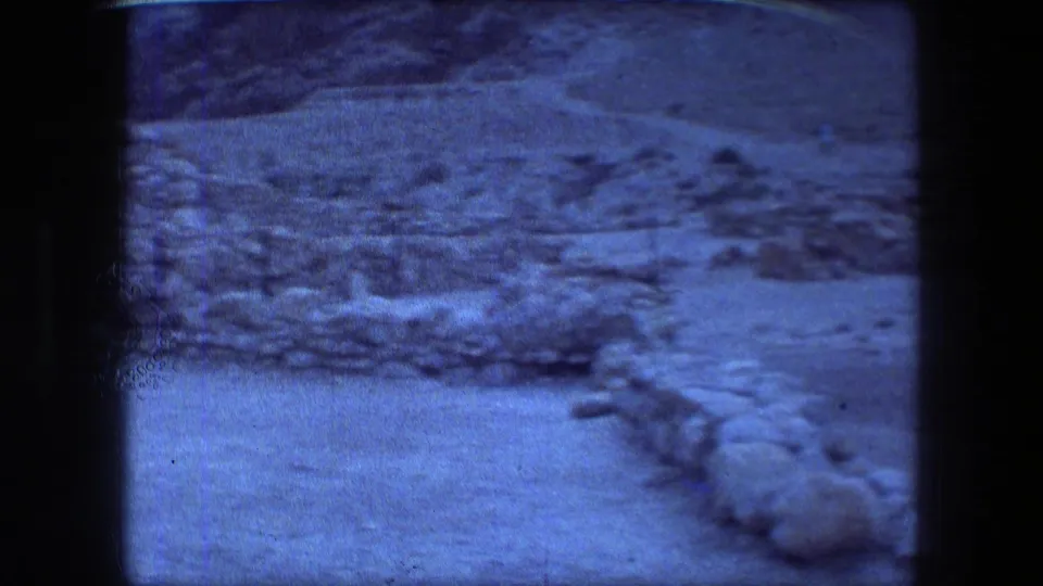 Arid Messada landscape with rocky ground, low stone edges, mountain slope, and the cool blue cast of the 8mm scan.