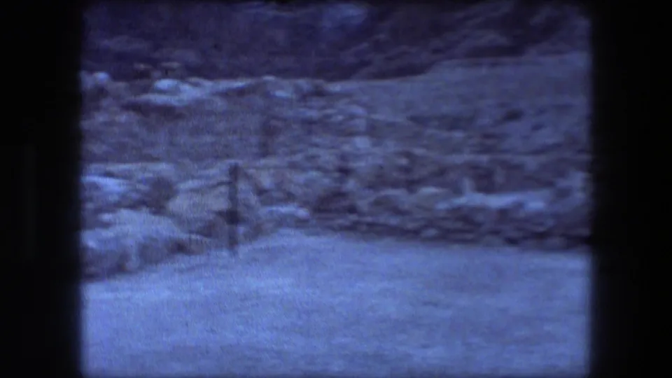 1976 8mm view of Masada stone ruins, scrubby desert ground, and distant rock slope with a blue-tinted home-movie look.
