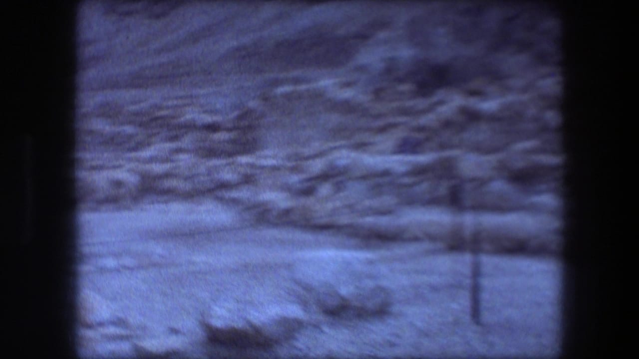 Blue-tinted 8mm frame of Masada's rocky desert terrain, low ruin lines, sparse shrubs, and a thin foreground fence post.