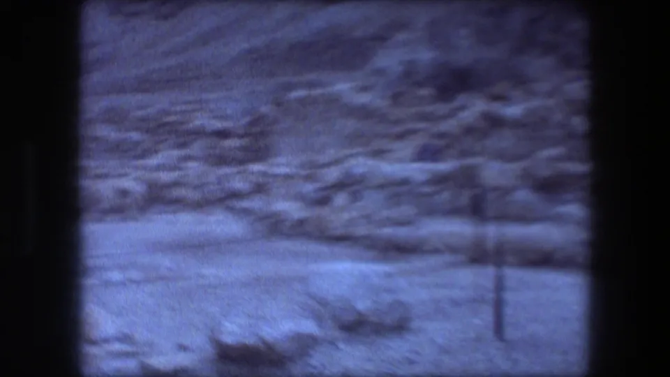 Blue-tinted 8mm frame of Masada's rocky desert terrain, low ruin lines, sparse shrubs, and a thin foreground fence post.