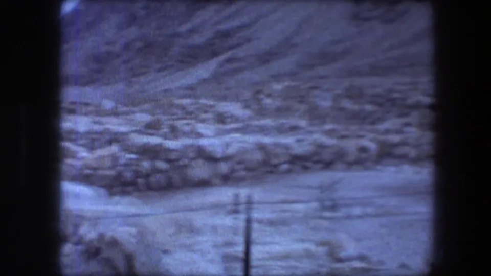 Handheld 1976 8mm view across Masada ruins, showing low stonework, rocky ground, scrub, and foreground wire fencing.