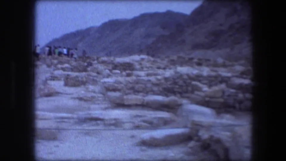 Wider 1976 8mm Masada landscape frame with low ruins, scattered rocks, sparse scrub, mountains, and hazy blue sky.