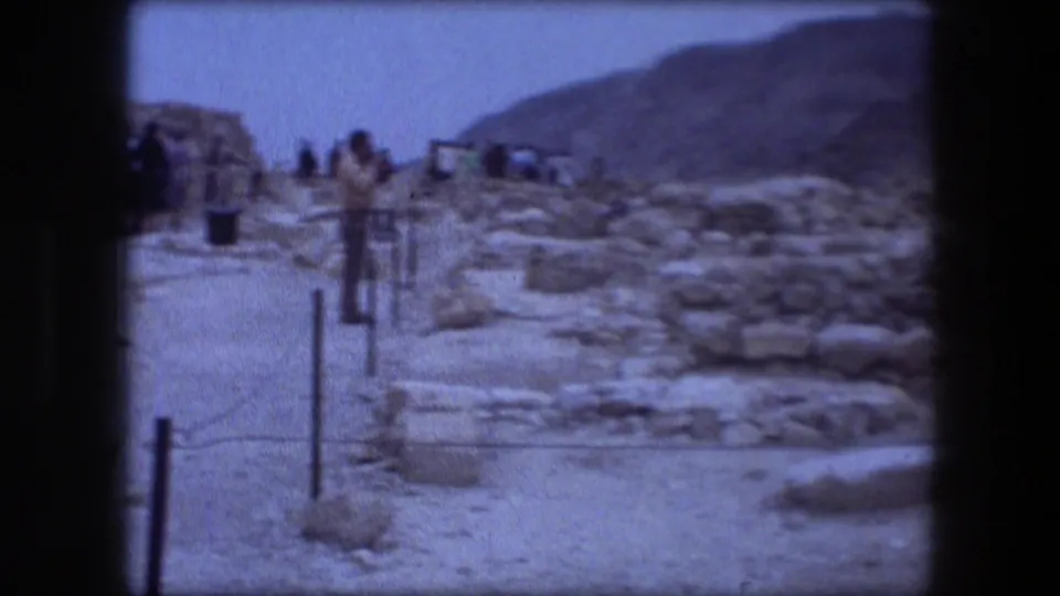 1976 8mm view of visitors at Messada, Israel, with stone ruins, rope barriers, dry ground, and hills beyond.