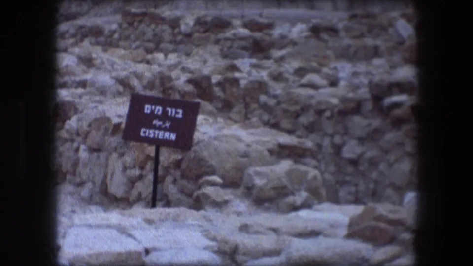 1976 8mm frame centers the bilingual cistern sign at Messada with rough stones, ruin walls, and pale desert ground.