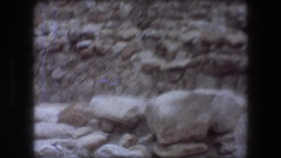 1976 8mm frame at Messada showing low stone remains, scattered rocks, and weathered blocks in soft focus.
