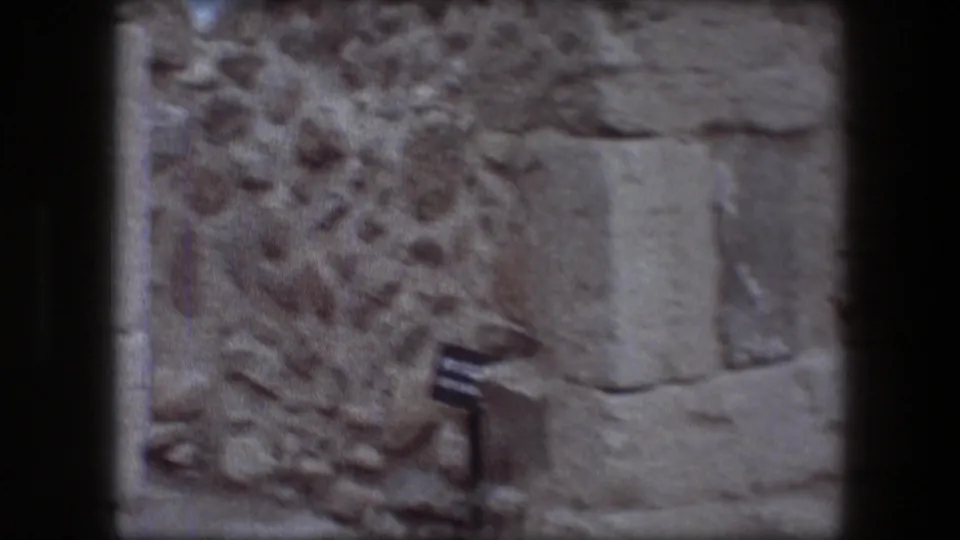 1976 8mm frame at Messada with a rough stone wall, rectangular carved slab, rubble, and a small site marker near the lower edge.