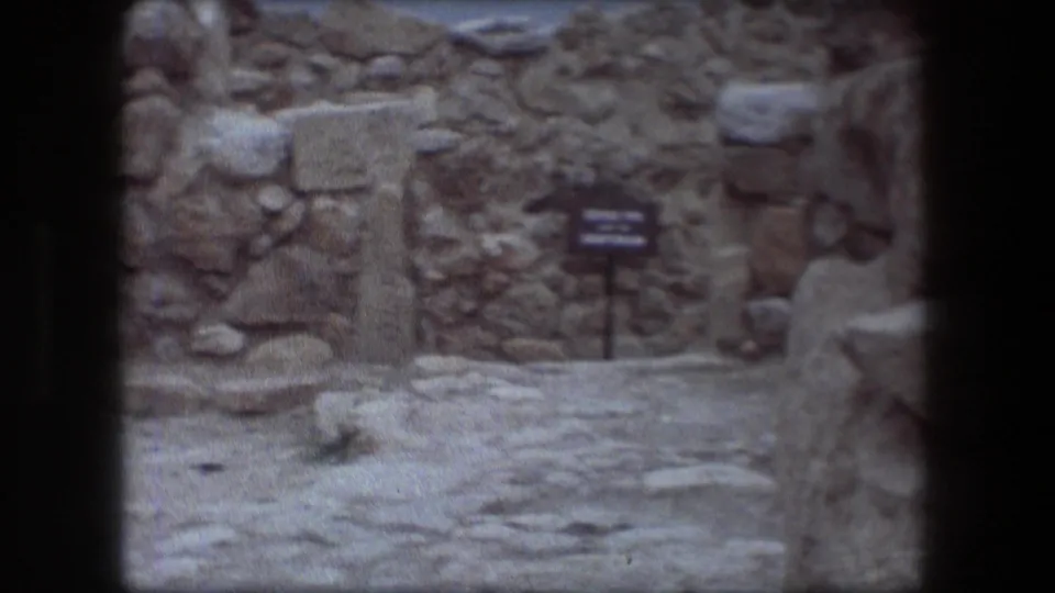 1976 Messada, Israel 8mm frame of a small sign, rough stone walls, and a pale stone path within the ruin site.