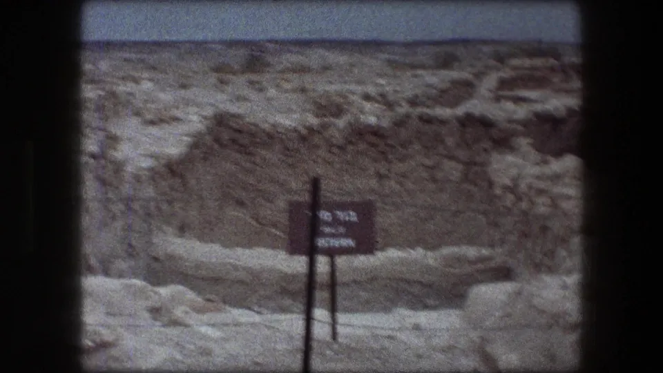 Messada, Israel ruins seen on 8mm film, with a dark bilingual-looking site sign standing before eroded stonework and desert ground.