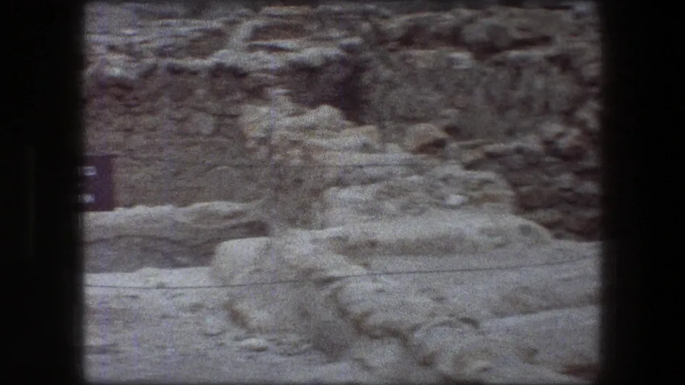Soft 8mm frame of Messada ruins, showing low stone walls, pale eroded rock and sandy ground with part of a sign at the left edge.