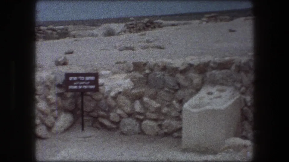 Visitor-shot 8mm view at Messada with a directional sign, rough stone walls, sandy terrain and a pale block marker in the foreground.