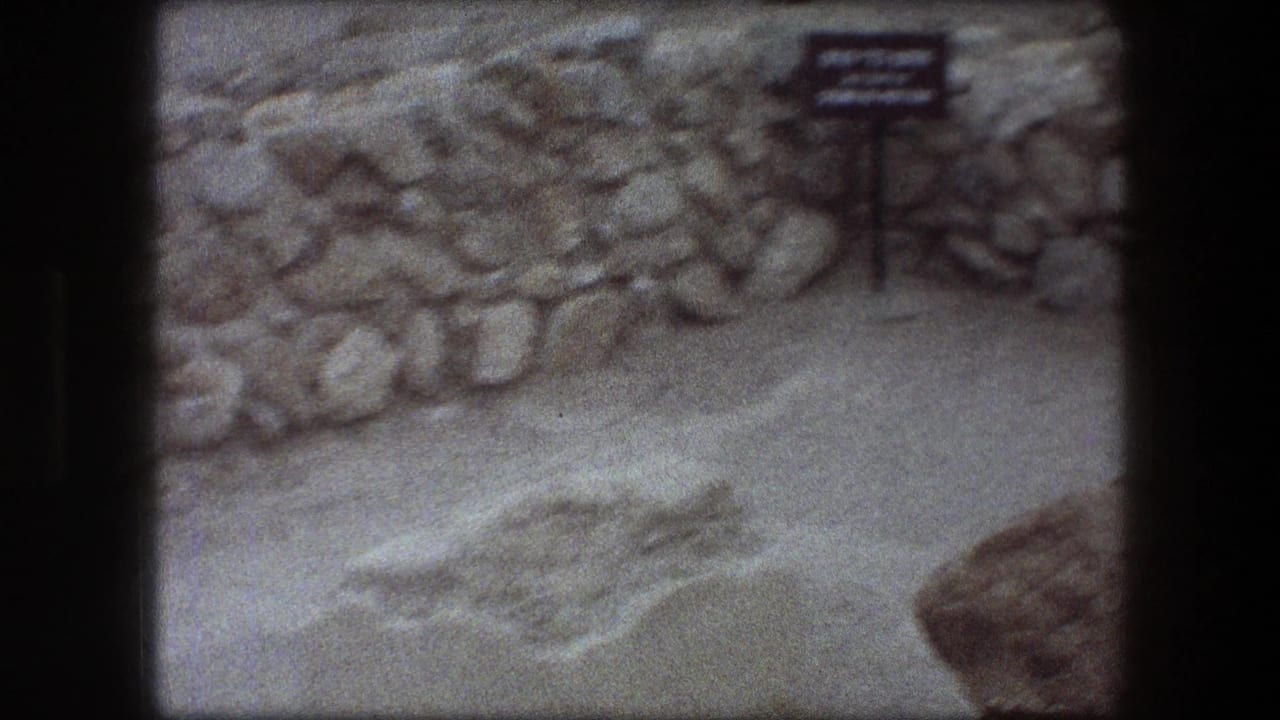 1976 Messada frame with a small bilingual site sign, stacked stones, pale sand, and dark 8mm frame borders.