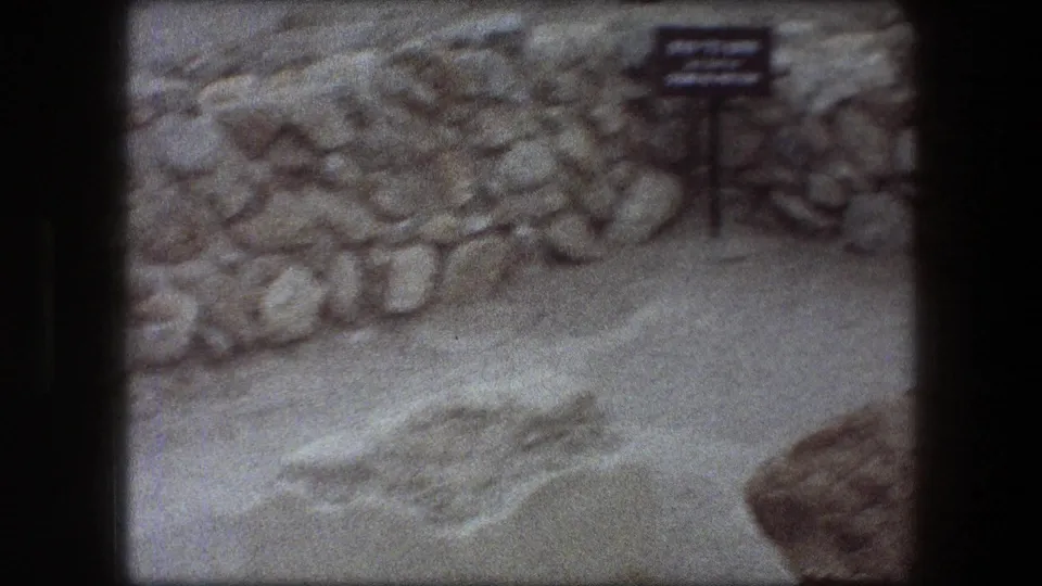 1976 Messada frame with a small bilingual site sign, stacked stones, pale sand, and dark 8mm frame borders.