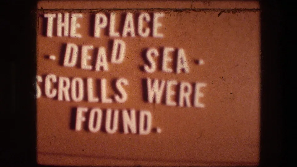 1976 8mm title-card frame with partial text about the place where the Dead Sea Scrolls were found beside cliff imagery.