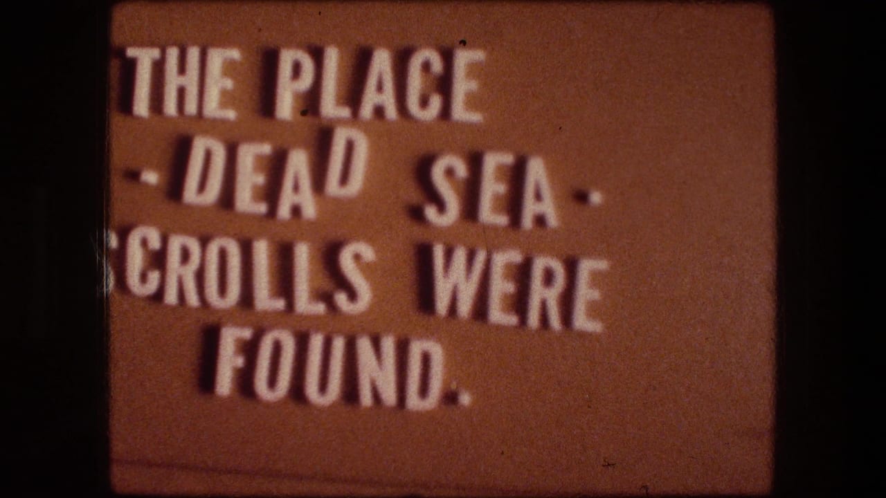 Warm-toned 8mm frame showing the same title card text about the Dead Sea Scrolls in soft focus.