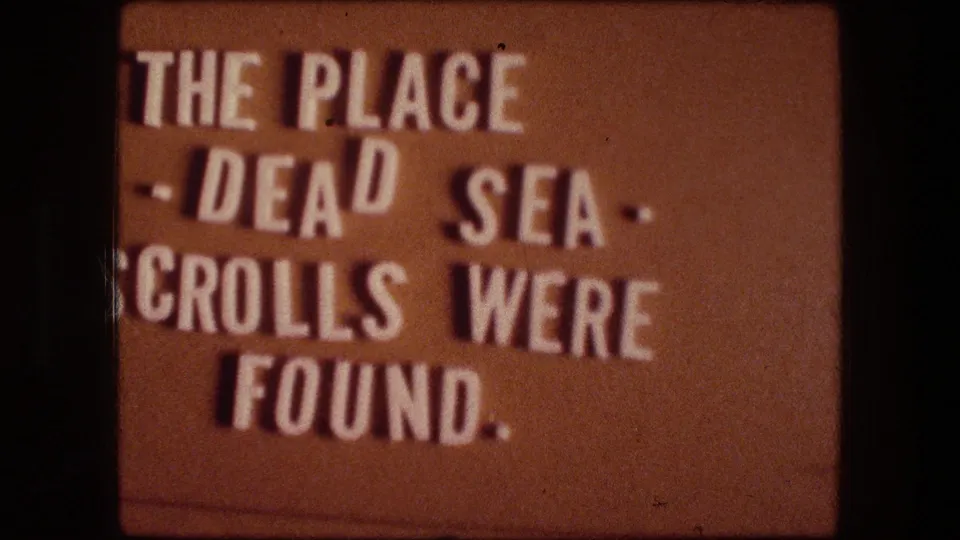 Warm-toned 8mm frame showing the same title card text about the Dead Sea Scrolls in soft focus.