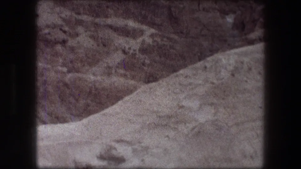 1976 8mm home-movie frame from Messada showing sandy foreground, layered arid slopes, and handheld framing within dark film borders.