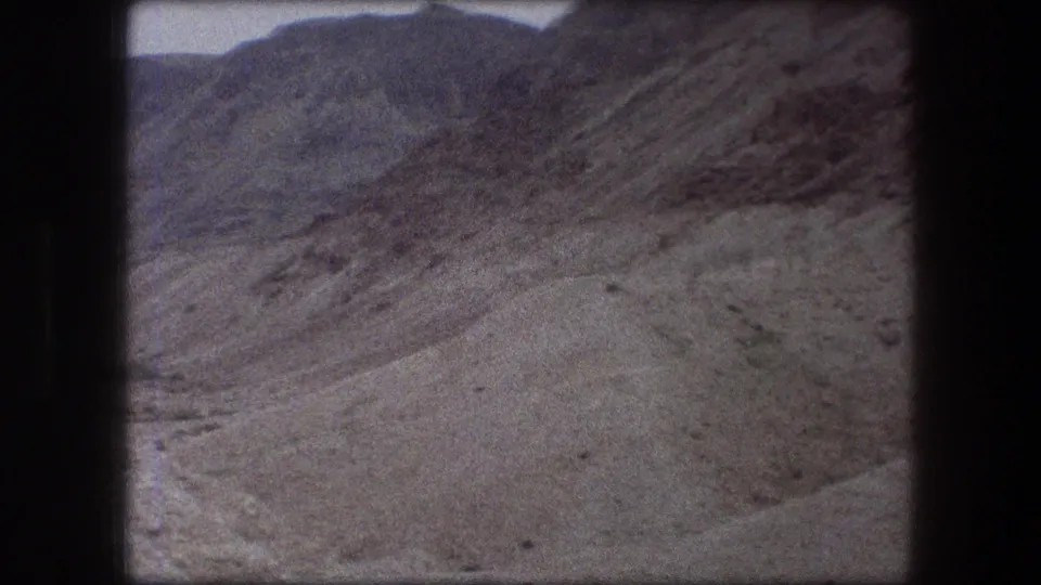 Grainy 8mm view of a broad Messada desert slope, with layered rock textures and sandy ground in the foreground.