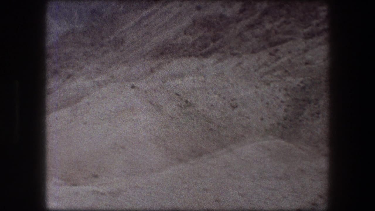 1976 Messada-area 8mm frame showing pale desert ground, low ridges, scattered rocks, and soft film grain.