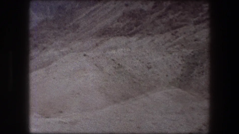 1976 Messada-area 8mm frame showing pale desert ground, low ridges, scattered rocks, and soft film grain.