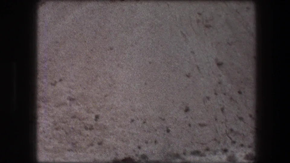 1976 8mm home-movie frame of Messada-area desert floor with pale sand, scattered stones, and subtle surface marks.
