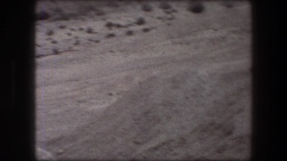 Messada-area 1976 8mm frame showing a wide sandy desert surface with scattered rocks, low ridges, and muted archival grain.