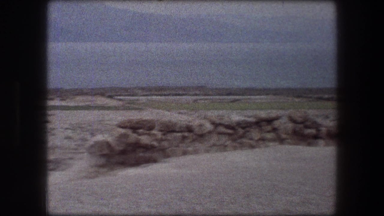 1976 Messada, Israel travel footage showing sandy ground, scattered rocks, a low wall, water, and distant mountains under a hazy sky.