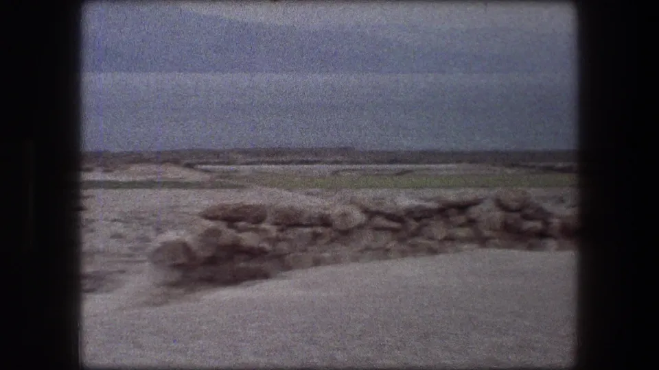1976 Messada, Israel travel footage showing sandy ground, scattered rocks, a low wall, water, and distant mountains under a hazy sky.