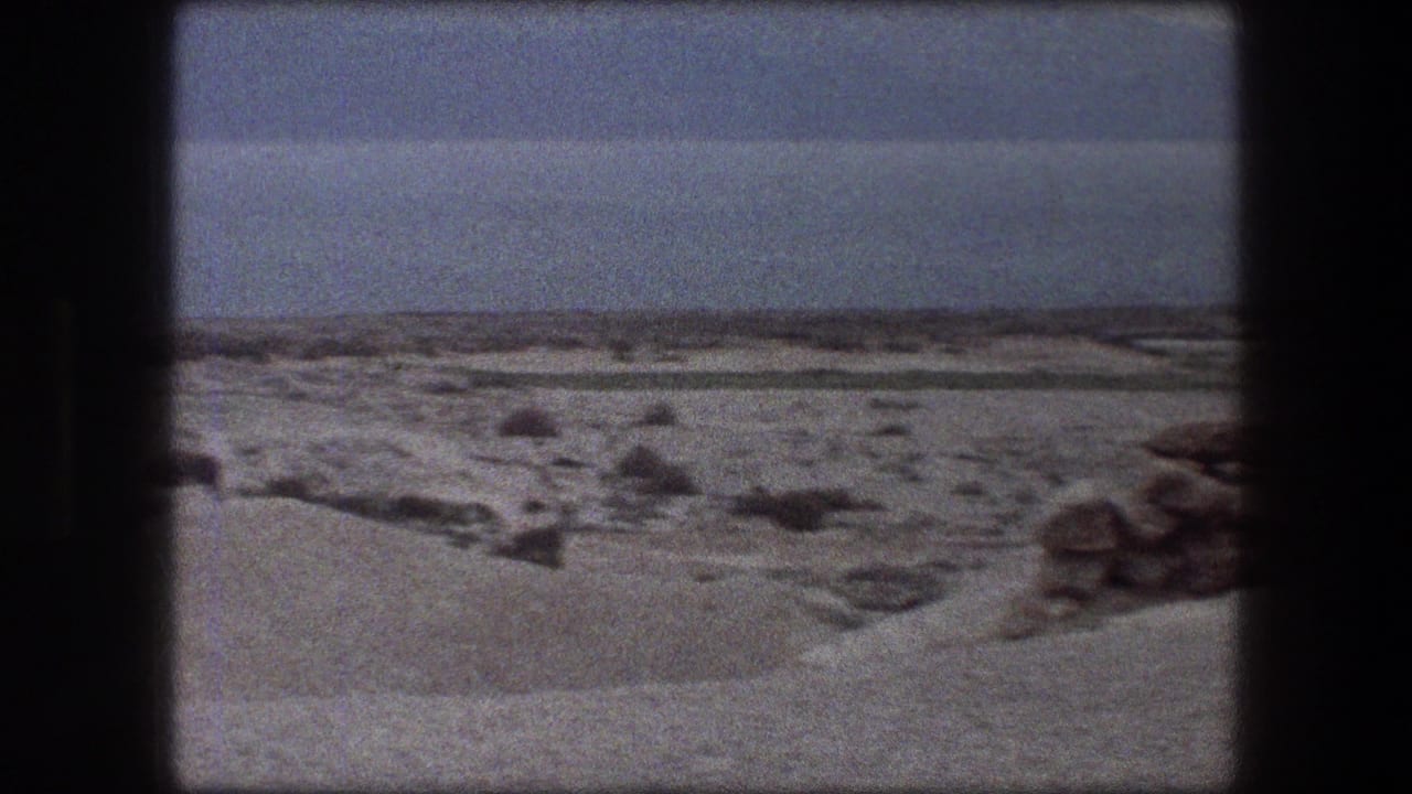1976 Messada, Israel home-movie frame with rocky desert ground, pale sand, water, and low mountains across the horizon.