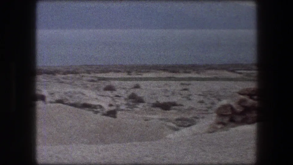 1976 Messada, Israel home-movie frame with rocky desert ground, pale sand, water, and low mountains across the horizon.