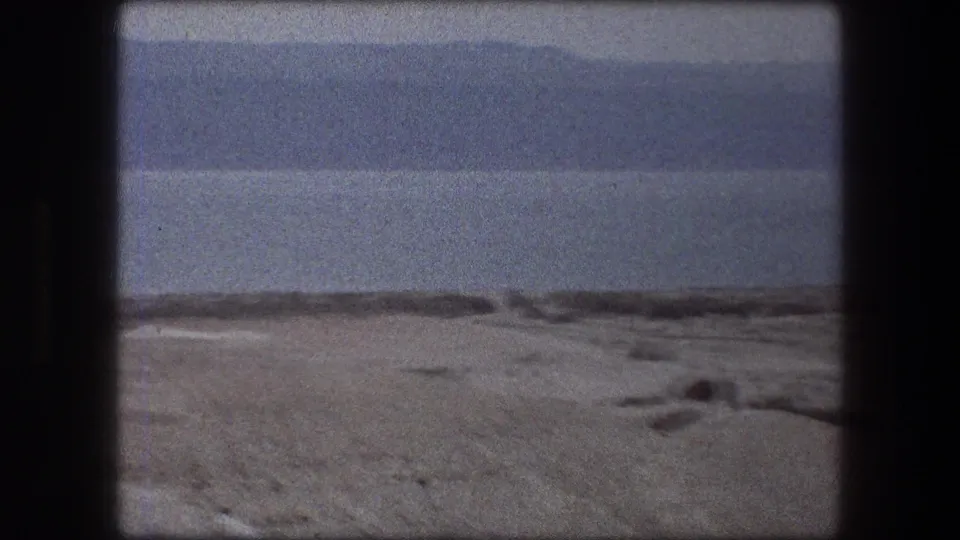 1976 Messada, Israel frame showing open desert ground, a dark horizontal ridge or shoreline, broad water, and hazy mountains.
