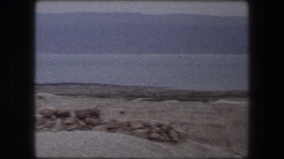 1976 Messada, Israel travel frame with a low stone wall, sandy ground, broad water, and muted mountains across the horizon.