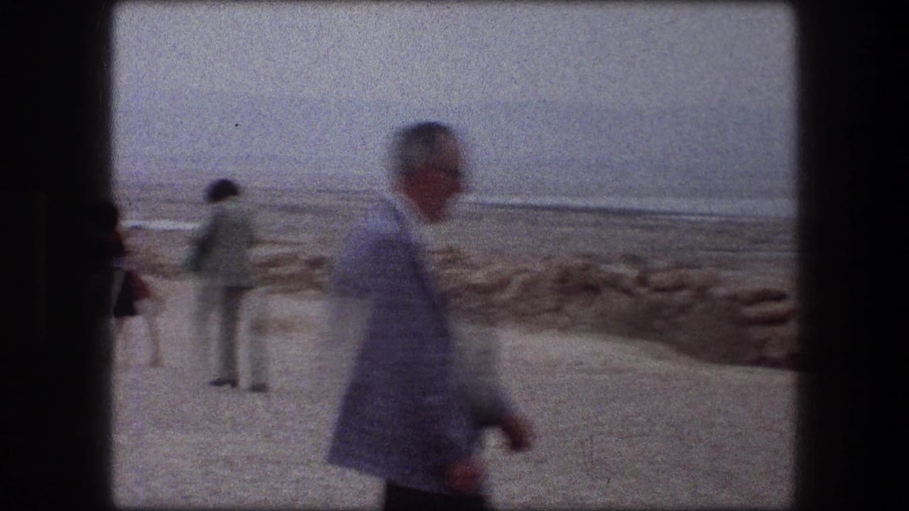 Man in a light jacket walks across a sandy beach area with other visitors near the water.
