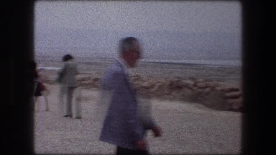 1976 Messada, Israel beach frame with a man crossing the foreground and visitors standing near the stone-lined shoreline.