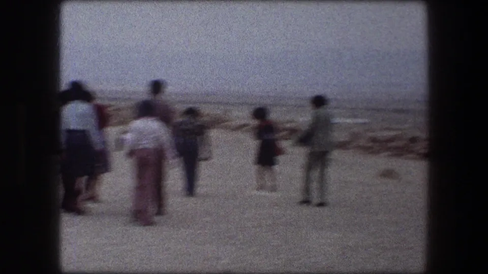 1976 8mm beach scene in Messada, Israel with several adults gathered on sand beside a low stone shoreline.